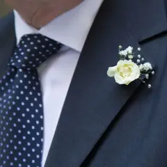 Elegancka butonierka co to? To ozdoba z białej róży i gipsówki, przypięta do ciemnej marynarki z kropkowanym krawatem.