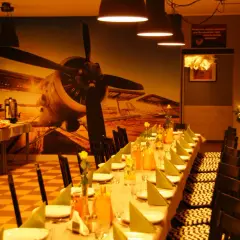Stół nakryty na kawalerski, z butelkami napojów i talerzami. W tle mural z samolotem.