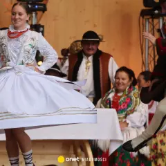 Zespoły góralskie w barwnych strojach tańczą na scenie. W centrum młoda kobieta w białej sukni, obok mężczyzna w brązowej sukmanie.