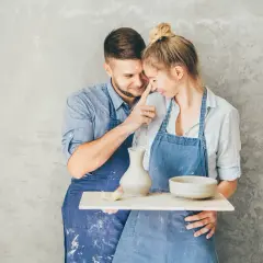 Para świętuje 9 rocznicę ślubu, tworząc ceramikę. Mężczyzna delikatnie dotyka nosa kobiety, oboje ubrani w fartuchy, z gliną na twarzach.