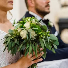 Panna młoda trzyma bukiet ślubny z białymi różami i zielenią, obok pan młody w garniturze.