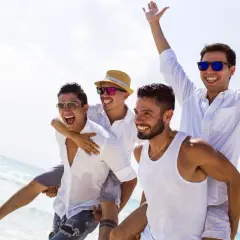 Czterech roześmianych mężczyzn na plaży, jeden niesie drugiego na barana. Wyglądają na to, kto organizuje wieczór kawalerski, świętując na wakacjach.