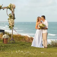 Para Młoda w objęciach, tuż obok kwiecistego łuku, celebruje swój ślub na plaży. W tle błękitne morze i palmy.