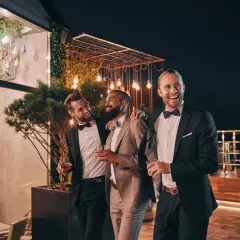 Trzech mężczyzn w eleganckich garniturach, z drinkami w rękach, śmieje się i rozmawia na tarasie. Jeden z nich, jako drużba, opiera rękę na ramieniu przyjaciela.