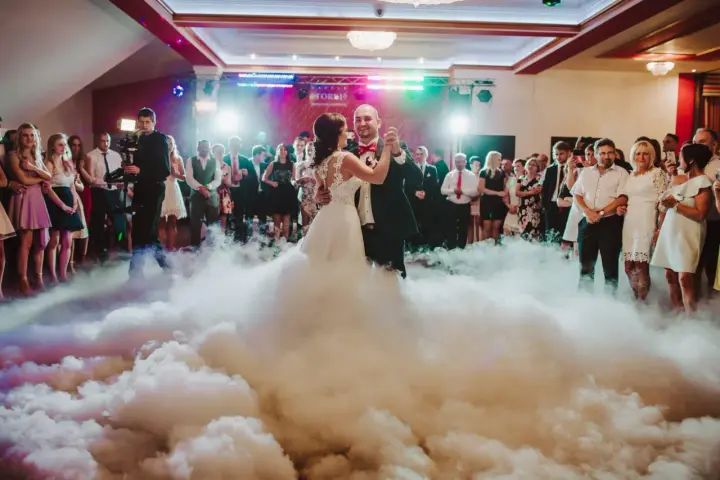 Para młoda tańczy w chmurach dymu na weselu, otoczona gośćmi.