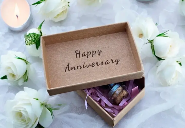 Otwarta szkatułka z napisem "Happy Anniversary", otoczona białymi różami i płonącą świecą. W środku mała buteleczka z korkiem.