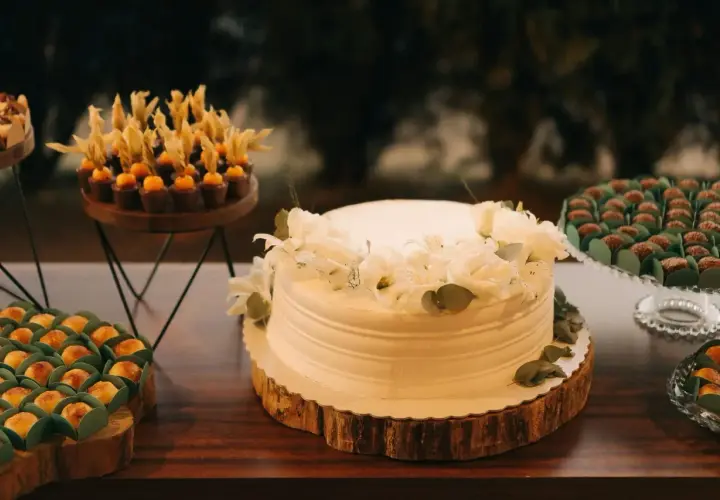 Biały tort komunijny ozdobiony białymi kwiatami i zielonymi liśćmi. Obok słodkości, które mogą być inspiracją, jakie kwiaty na tort komunijny.