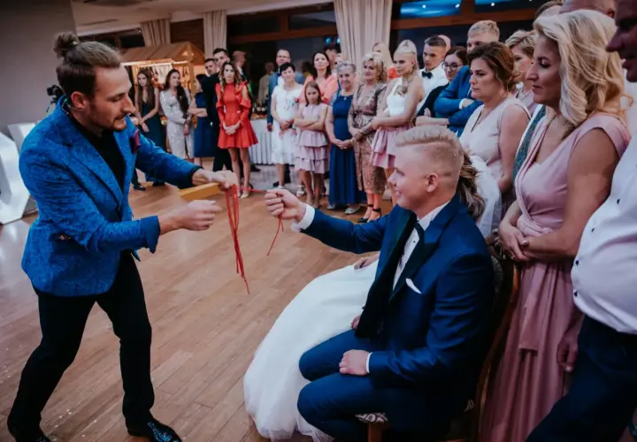 Iluzjonista na wesele wykonuje sztuczkę z czerwonymi wstążkami dla pana młodego.