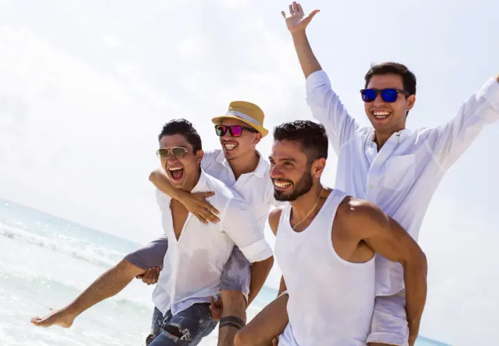 Czterech roześmianych mężczyzn na plaży, jeden niesie drugiego na barana. Wyglądają na to, kto organizuje wieczór kawalerski, świętując na wakacjach.