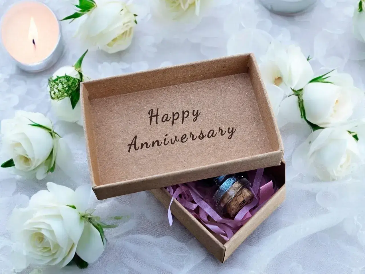 Otwarta szkatułka z napisem "Happy Anniversary", otoczona białymi różami i płonącą świecą. W środku mała buteleczka z korkiem.