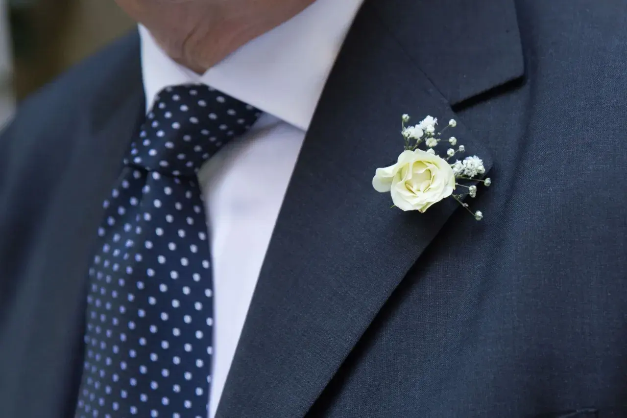 Elegancka butonierka co to? To ozdoba z białej róży i gipsówki, przypięta do ciemnej marynarki z kropkowanym krawatem.
