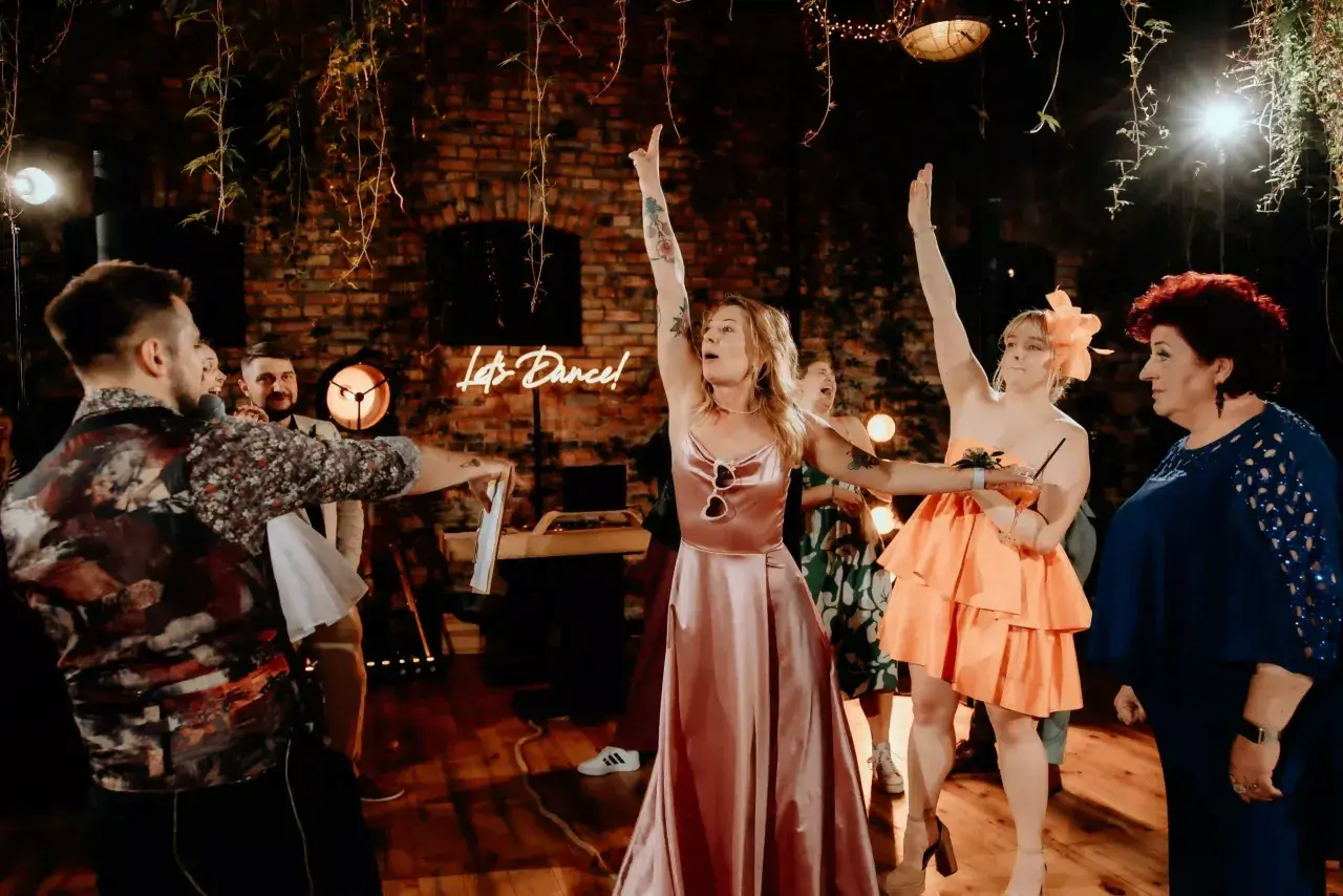 Nowoczesne oczepiny w rustykalnym klimacie. Kobiety w sukniach unoszą ręce, a neonowy napis "Let's Dance!" zaprasza do zabawy.
