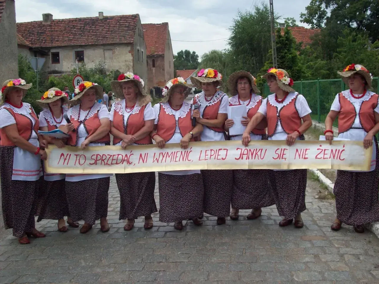 Kobiety w ludowych strojach trzymają transparent z napisem "Ni to sprzedać, ni wymienić, lepiej Janku się nie żenić". To śmieszne teksty na bramy weselne.