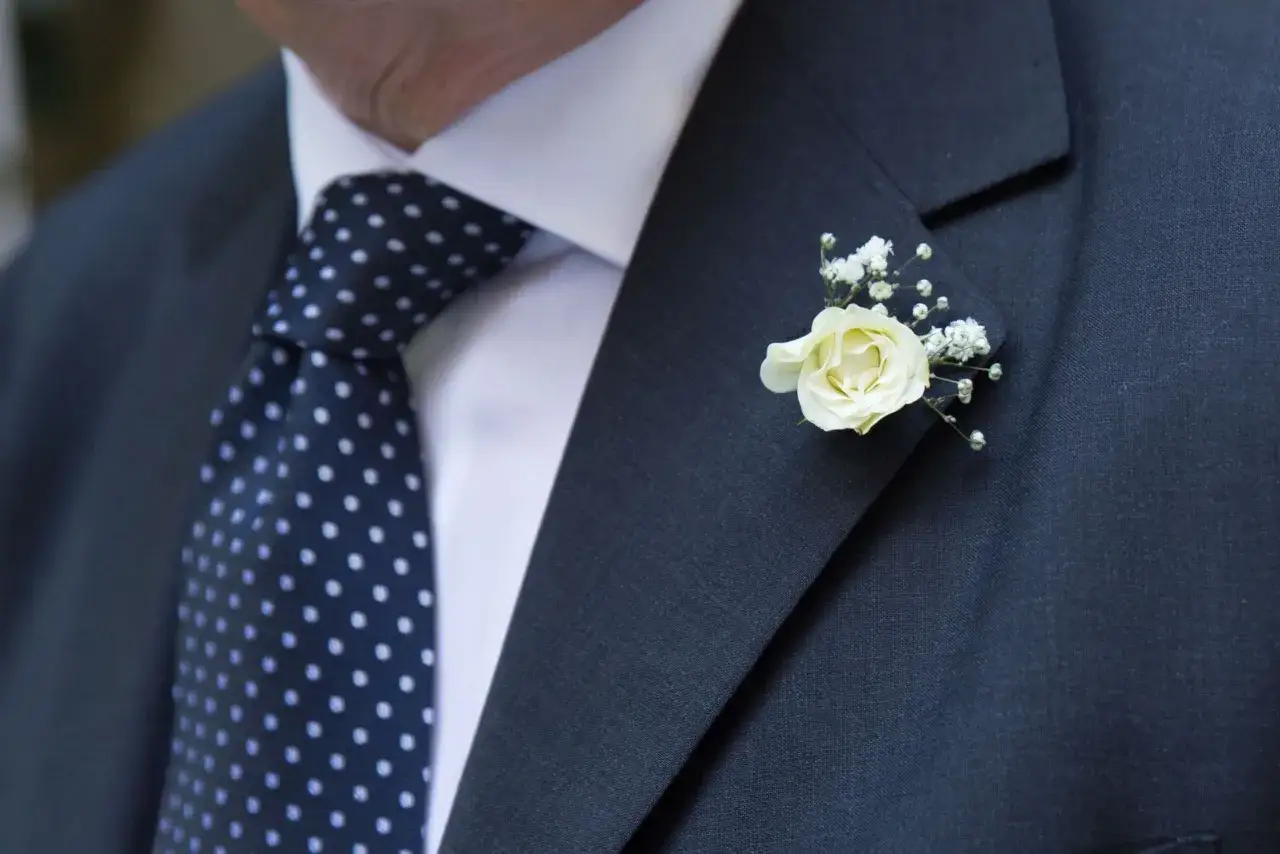 Elegancka męska stylizacja: granatowy garnitur, biała koszula, kropkowany krawat i biała róża jako butonierka.