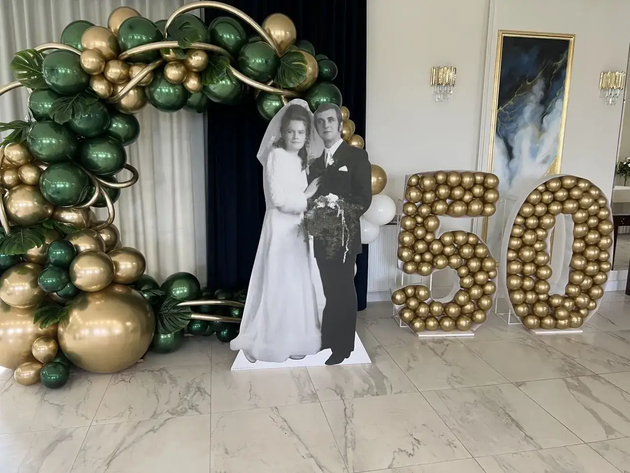 Dekoracja na 50 rocznicę ślubu: para młoda na zdjęciu, balony w kształcie cyfr 50 i łuk z zielonych i złotych balonów.