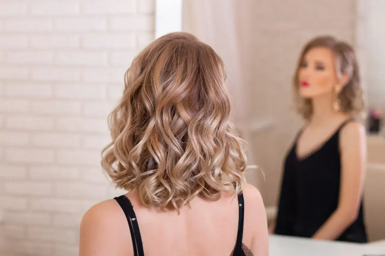 Eleganckie upięcia okolicznościowe dla blondynki z falami. W lustrze widać jej makijaż i biżuterię.