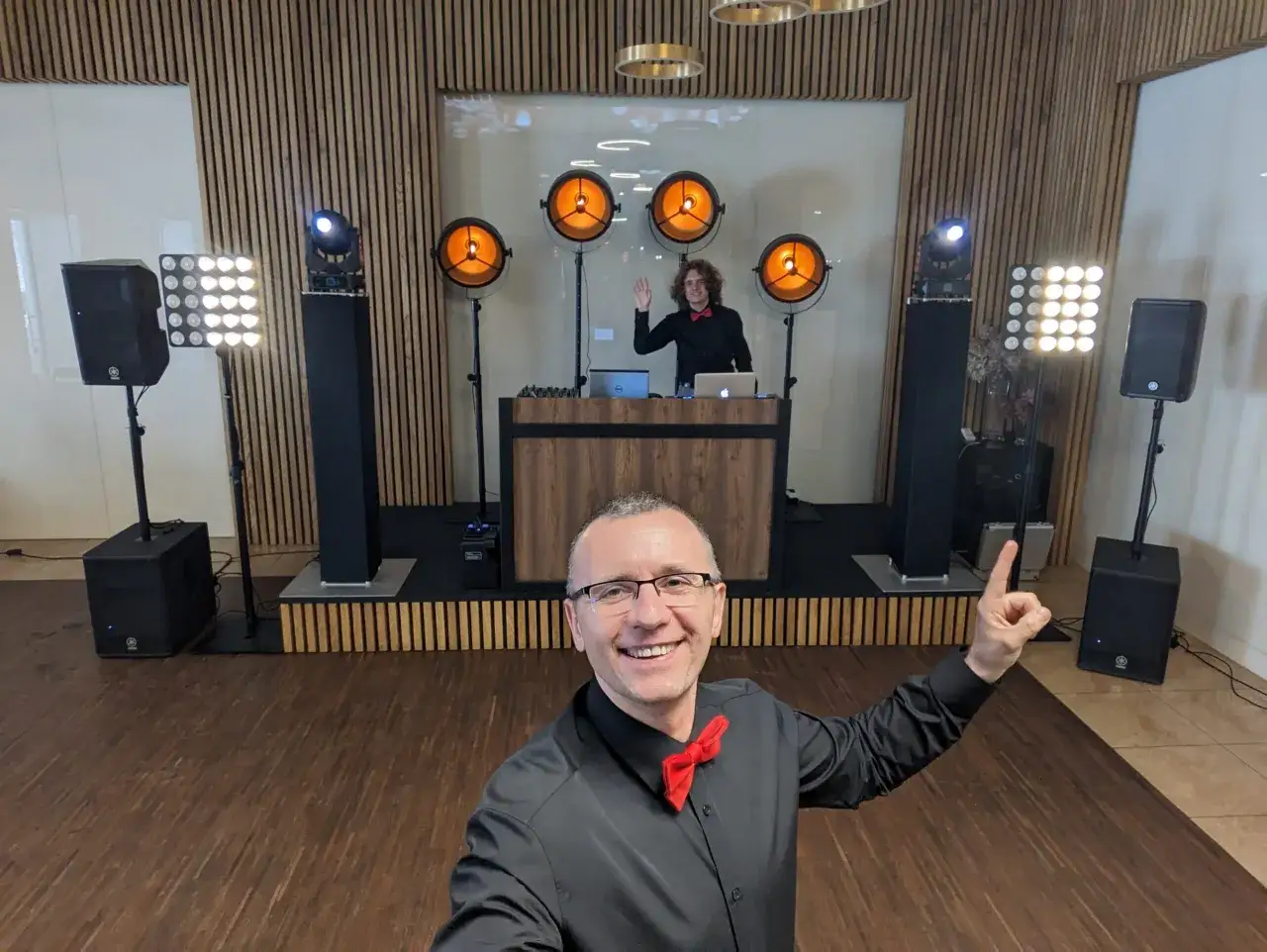 Uśmiechnięty dj na weselu, gotowy do zabawy. W tle sprzęt nagłośnieniowy i oświetlenie.